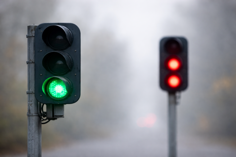 Deux feux de signalisation autonomes : l’un affiche un signal vert net au premier plan, l’autre un double signal rouge flou à distance, dans un environnement neutre et brumeux.